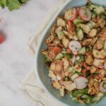 Grilled chicken salad with radishes, cucumbers, and fresh herbs in a white bowl on a cozy cloth, surrounded by radishes on marble surface, perfect for healthy recipes and summer meals.