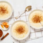 Creamy rice pudding garnished with cinnamon, served in three white bowls on a white wooden table with spices like cinnamon sticks and orange zest for flavor.
