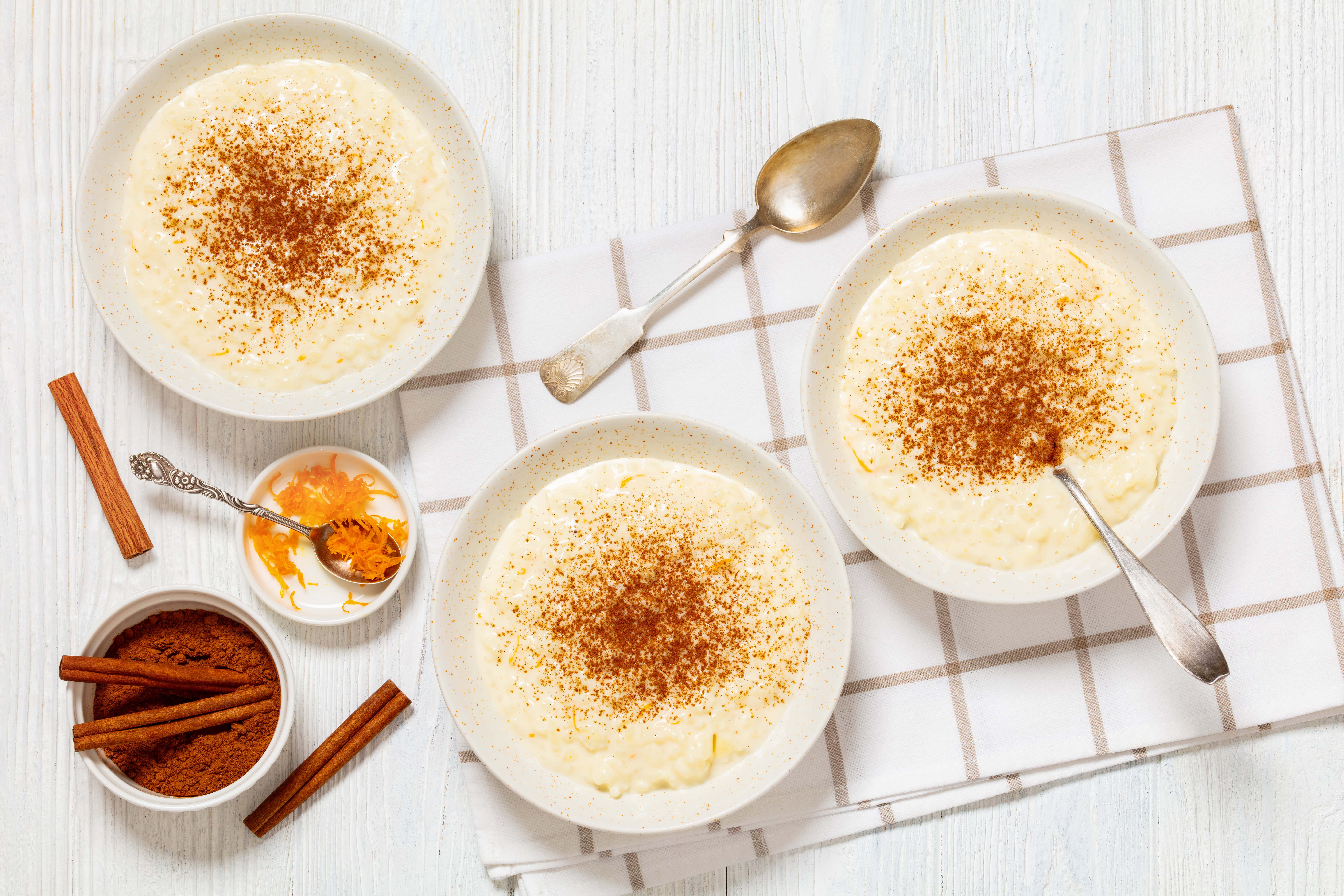 Creamy rice pudding garnished with cinnamon, served in three white bowls on a white wooden table with spices like cinnamon sticks and orange zest for flavor.