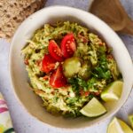 Creamy guacamole with fresh tomatoes, chopped cilantro, lime wedges, jalapeño, served with crispy whole grain crackers, on a light gray textured surface.
