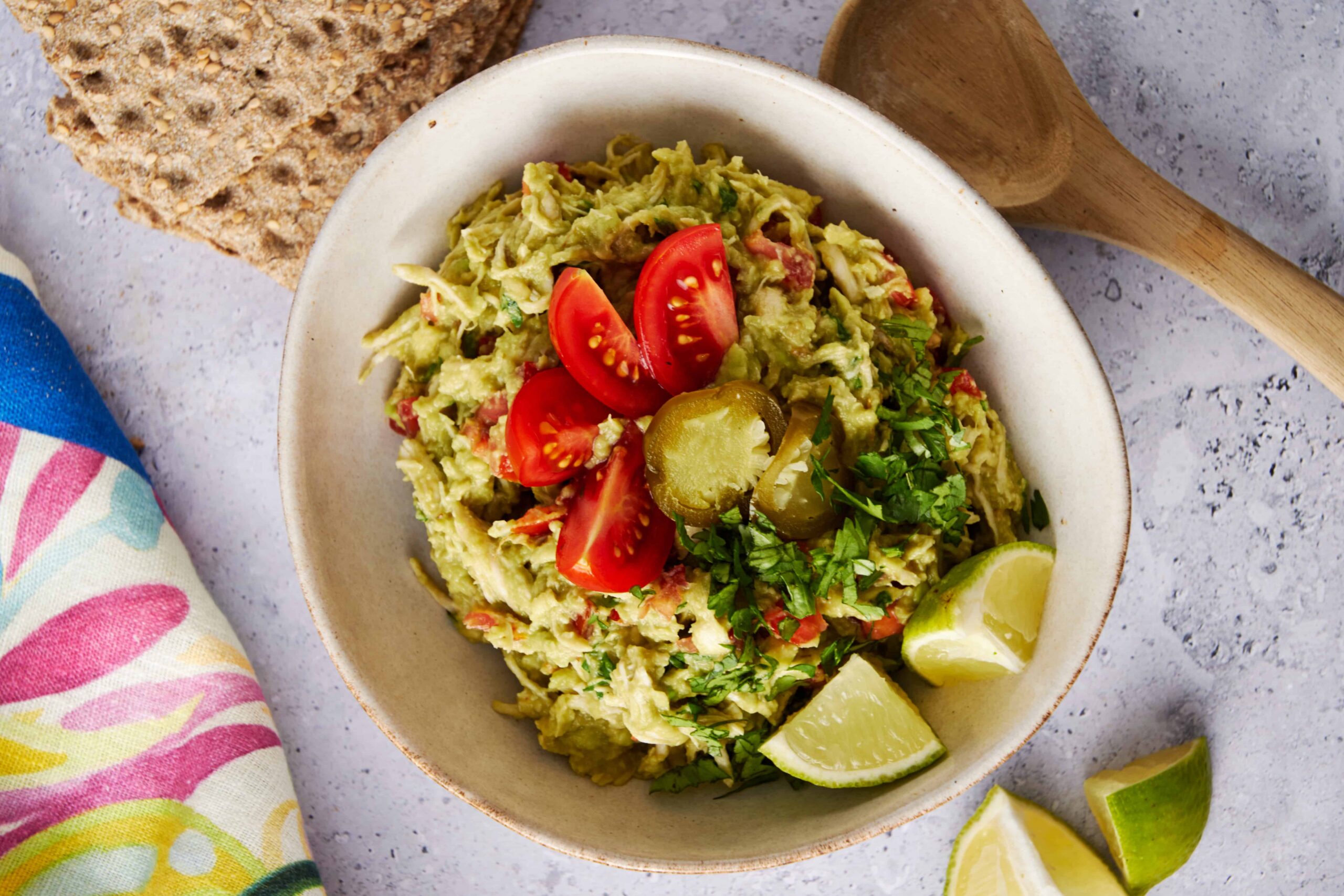 Creamy guacamole with fresh tomatoes, chopped cilantro, lime wedges, jalapeño, served with crispy whole grain crackers, on a light gray textured surface.
