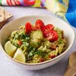 Creamy guacamole topped with cherry tomatoes, pickles, and fresh herbs served in a white bowl with lime wedges and crispy crackers nearby.