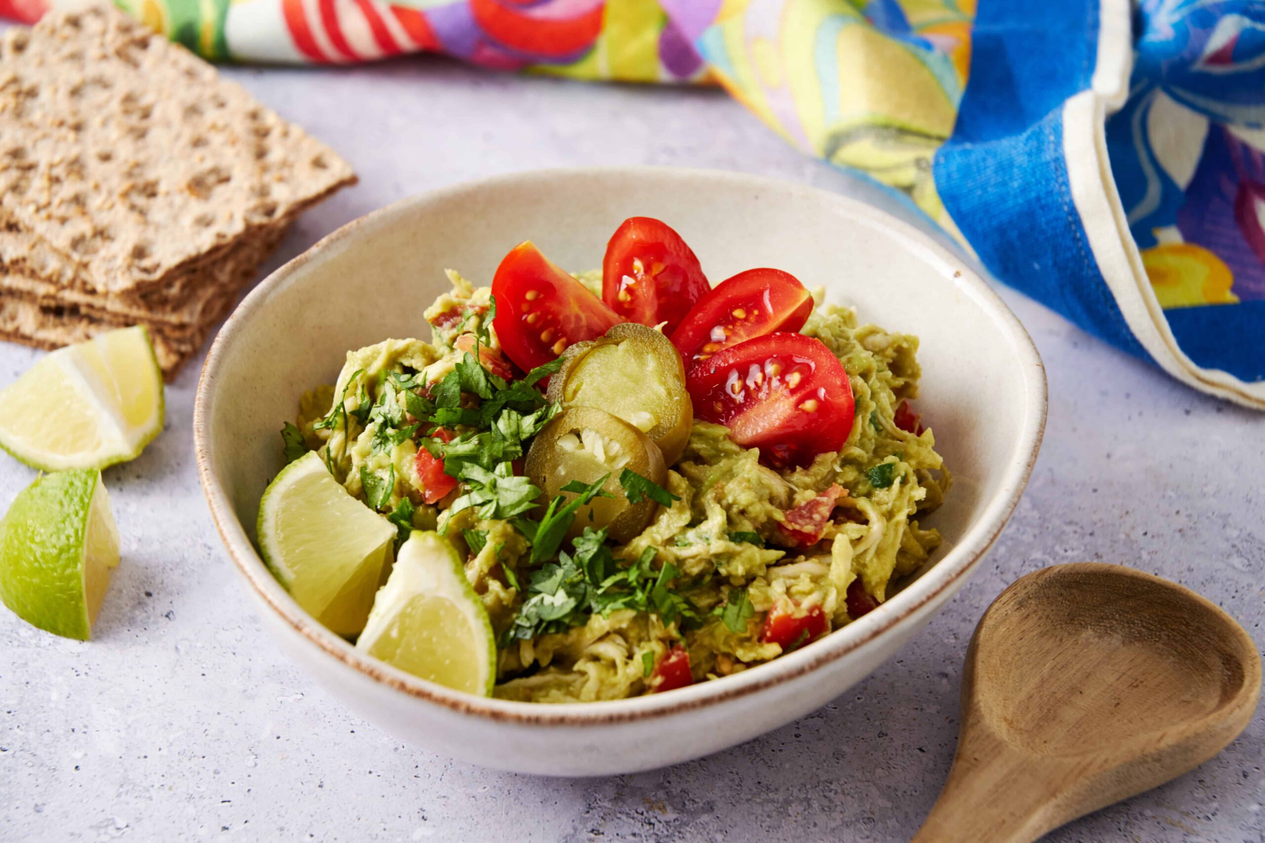 Creamy guacamole topped with cherry tomatoes, pickles, and fresh herbs served in a white bowl with lime wedges and crispy crackers nearby.