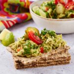 Creamy avocado and tomato spread on whole grain crispbread with fresh lime and cilantro garnish, served as a healthy snack or appetizer. Perfect for avocado lovers and nutritious snacks.