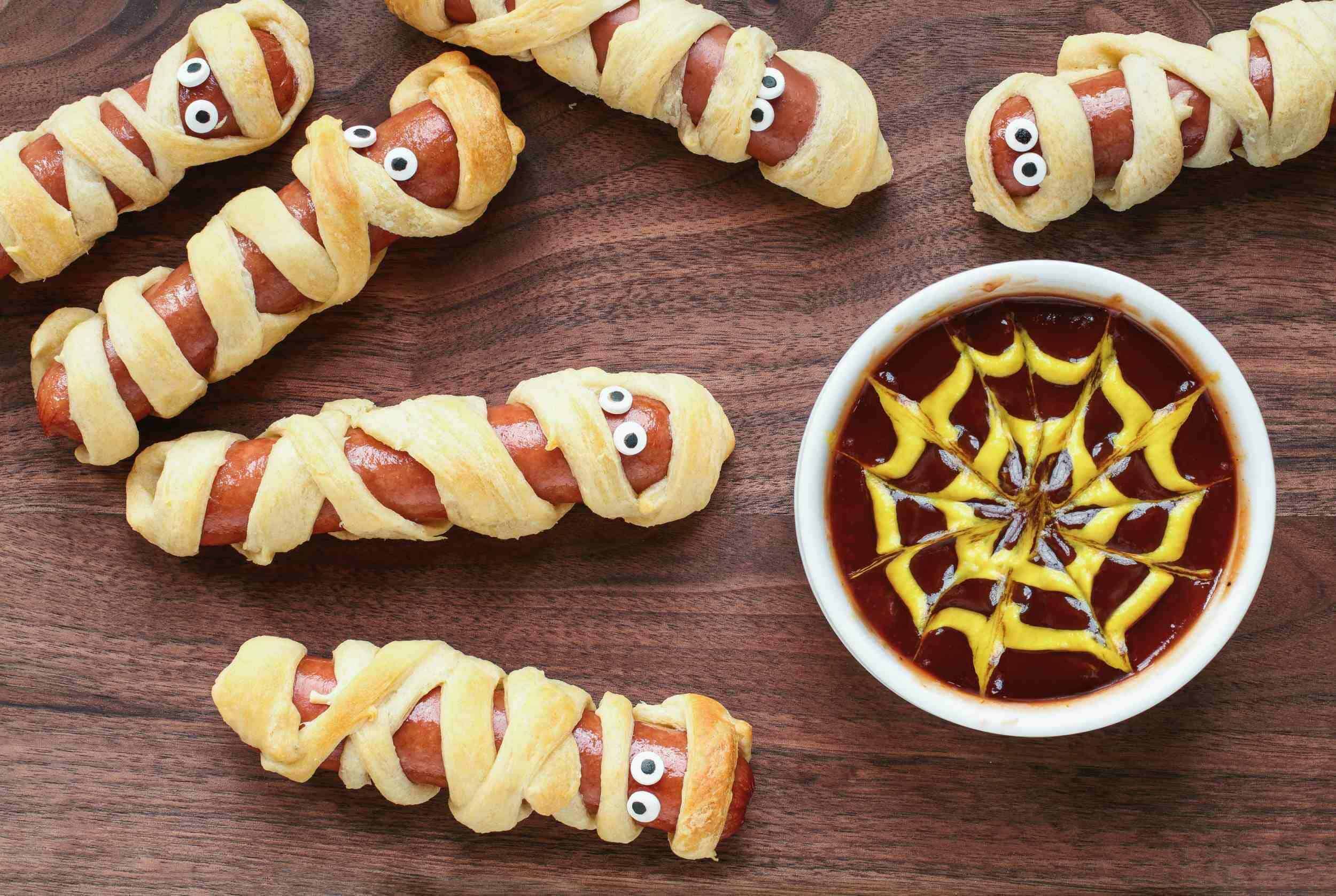 Creepy Halloween hot dogs wrapped in puff pastry with edible googly eyes, paired with a spooky spider web Halloween sauce on a wooden board. Perfect for a fun and festive party treat.