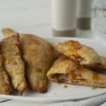 Crispy fried empanadas filled with ham and cheese on a white plate, served with a side of drinks and water in the background, perfect for a savory snack or meal.