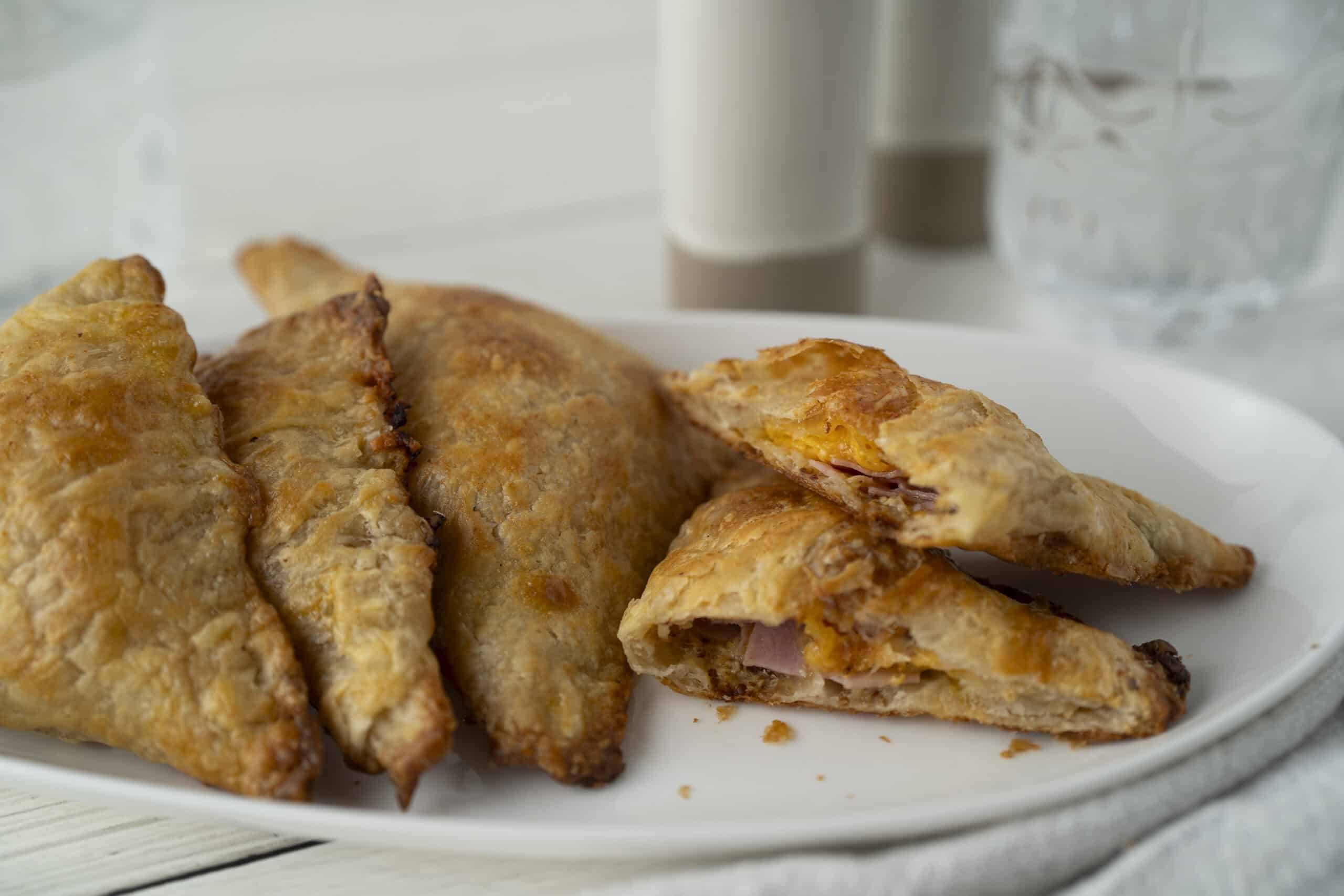 Crispy fried empanadas filled with ham and cheese on a white plate, served with a side of drinks and water in the background, perfect for a savory snack or meal.