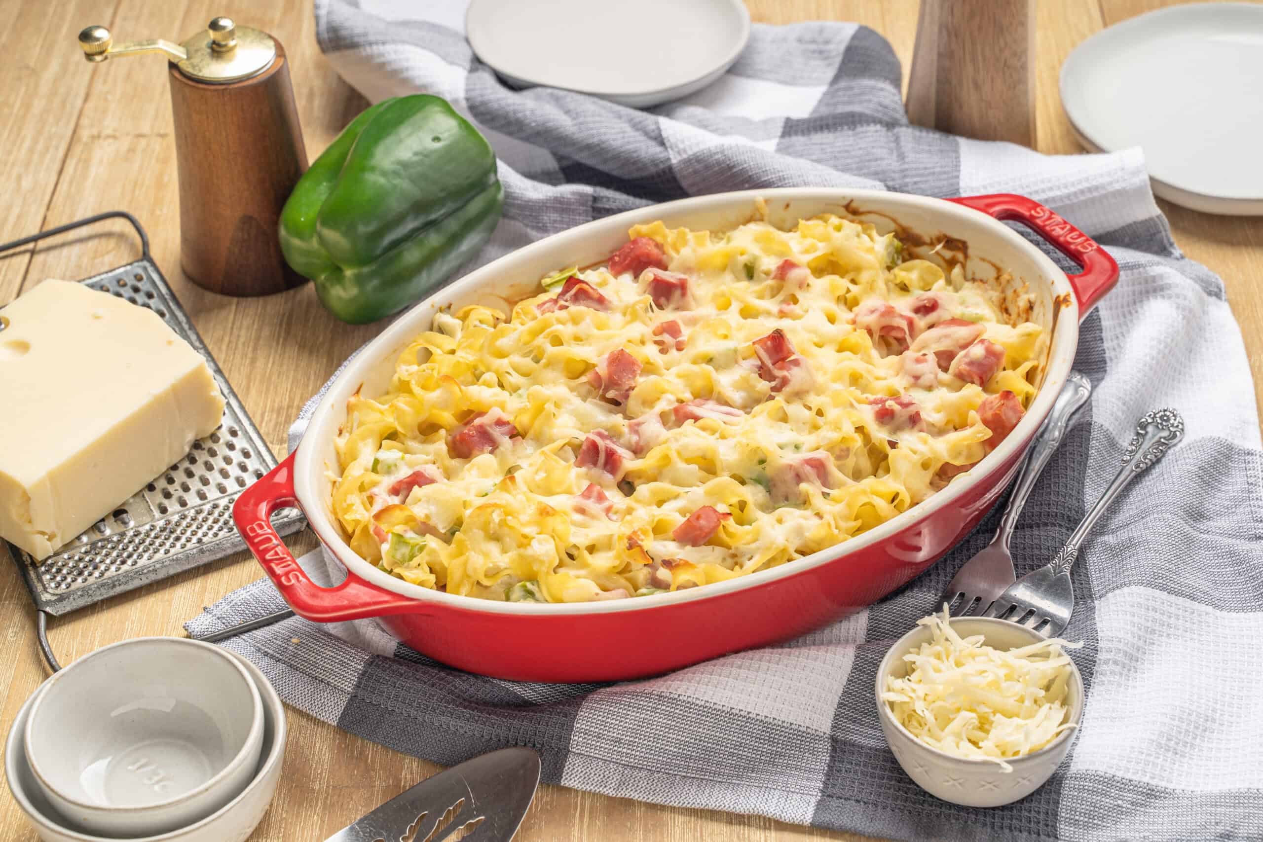Creamy pasta casserole with ham, cheese, and vegetables baked in a red oval dish, served with shredded cheese, pepper, and fresh green pepper on a rustic wooden table.