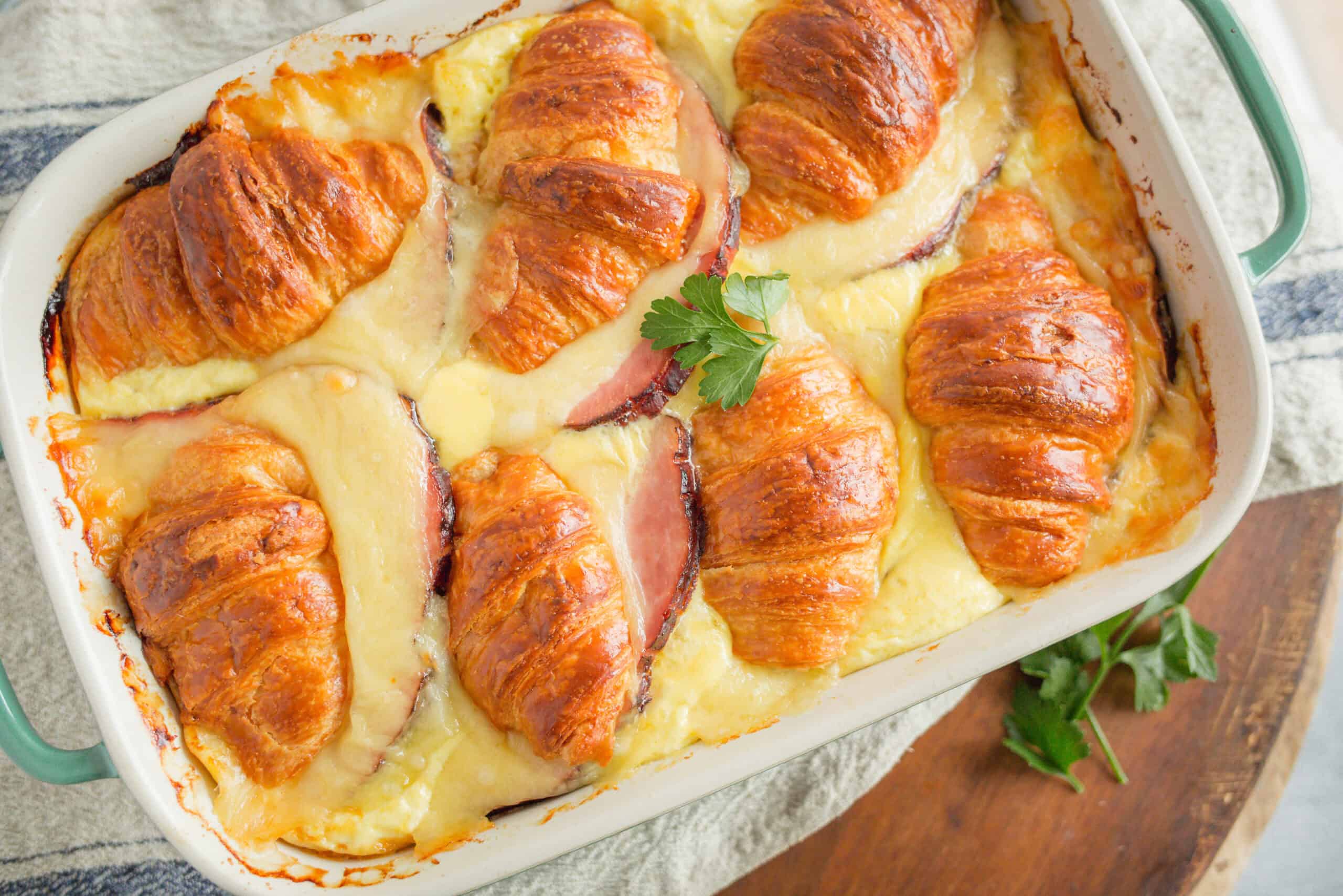 Golden-brown croissants baked with ham and melted cheese in a casserole dish, garnished with cilantro, for a delicious breakfast or brunch recipe.