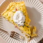 Creamy ham and cheese breakfast casserole topped with whipped cream on a white plate, served with silver forks, perfect for brunch or hearty breakfast.