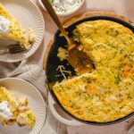 Creamy breakfast casserole with eggs, ham, cheese, and herbs served in a cast iron skillet, topped with fresh chives, with slices on white plates, sour cream garnished with chives on the side.
