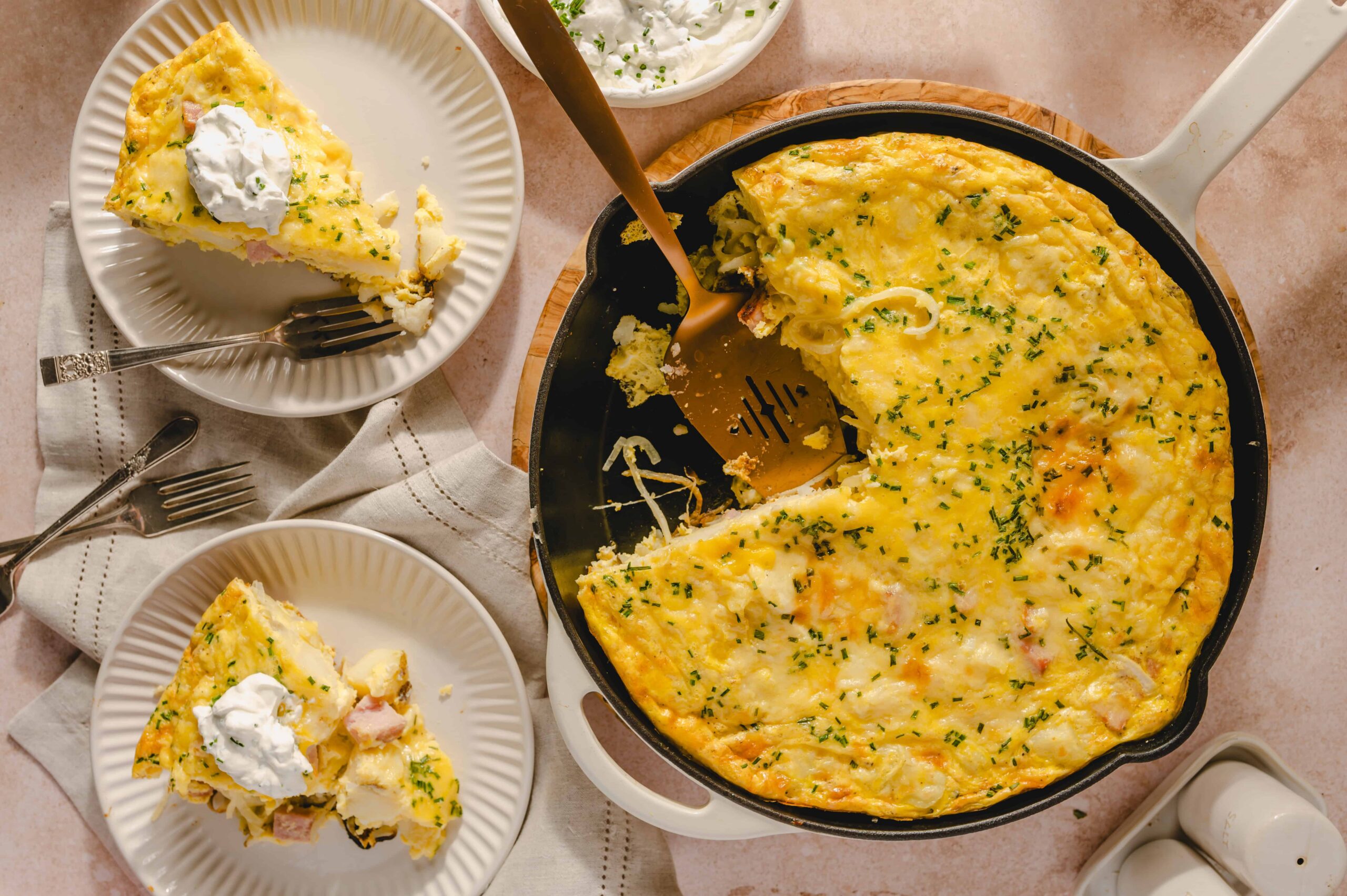 Creamy breakfast casserole with eggs, ham, cheese, and herbs served in a cast iron skillet, topped with fresh chives, with slices on white plates, sour cream garnished with chives on the side.