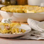 Creamy potato and ham casserole served with sour cream on a white plate, featuring a baked golden crust and melted cheese, perfect for comforting family meals.