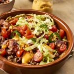 Savory loaded hash brown casserole topped with cherry tomatoes, jalapeƱos, shredded lettuce, bacon, and melted cheese in a rustic ceramic dish. Perfect for breakfast or brunch.