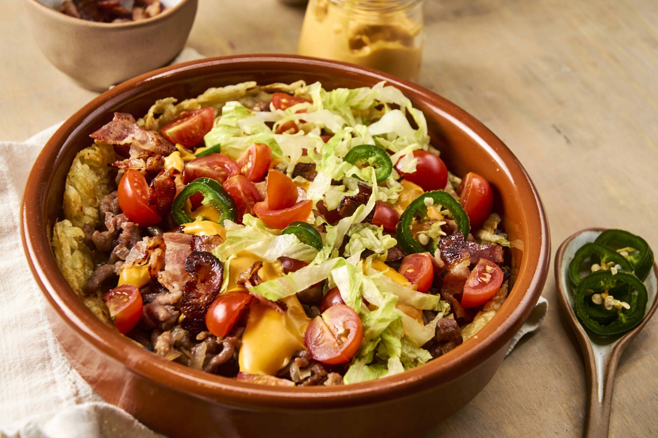 Savory loaded hash brown casserole topped with cherry tomatoes, jalapeƱos, shredded lettuce, bacon, and melted cheese in a rustic ceramic dish. Perfect for breakfast or brunch.