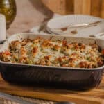 Baked cheesy casserole with vegetables in a black and white ceramic dish on a wooden cutting board.