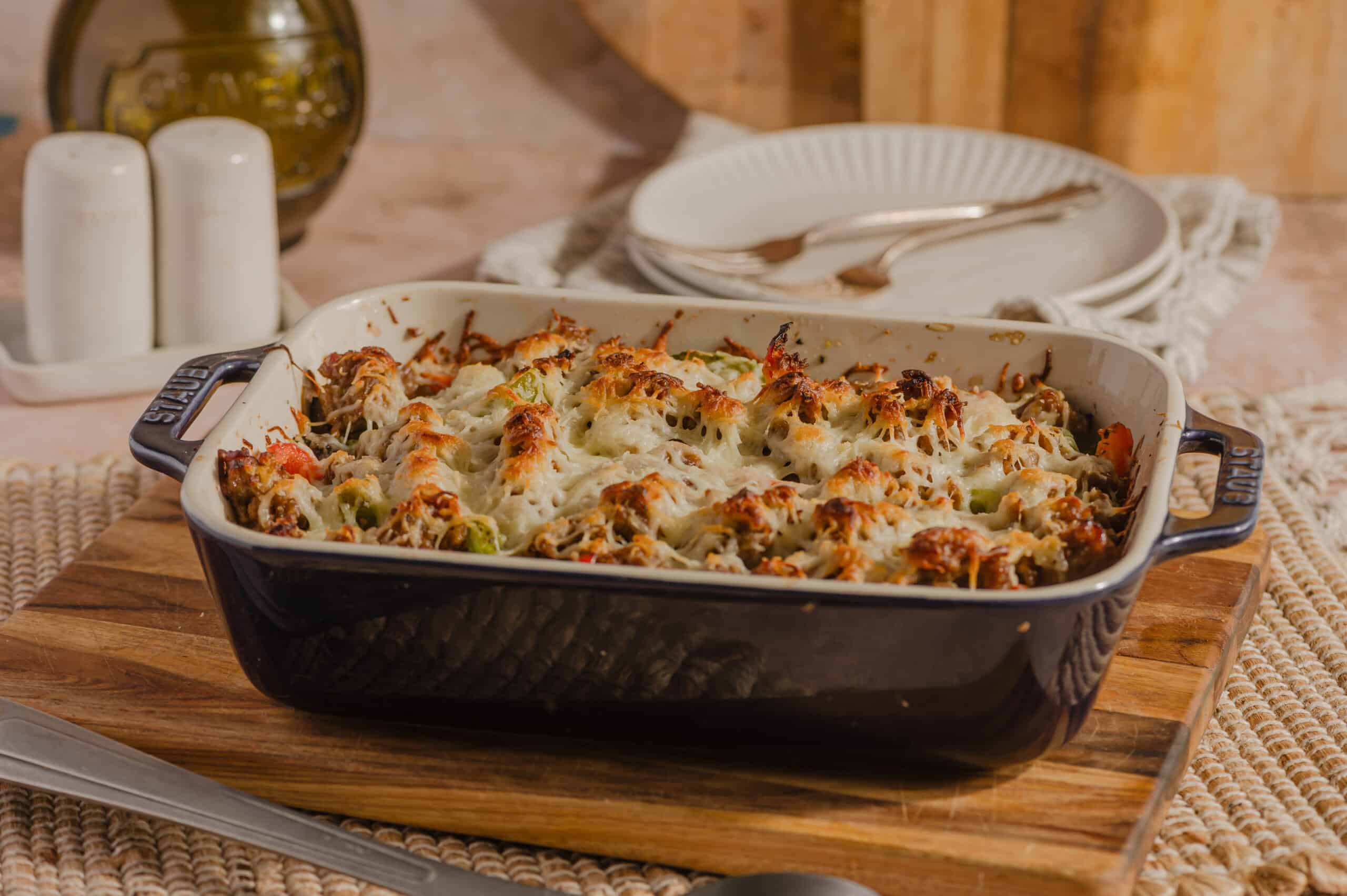 Baked cheesy casserole with vegetables in a black and white ceramic dish on a wooden cutting board.
