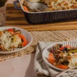 Cheesy lasagna baked dish with layers of melted cheese, seasoned ground meat, and fresh vegetables, served on white plates with colorful peppers and mushrooms, wooden cutting board and baking dish in the background.
