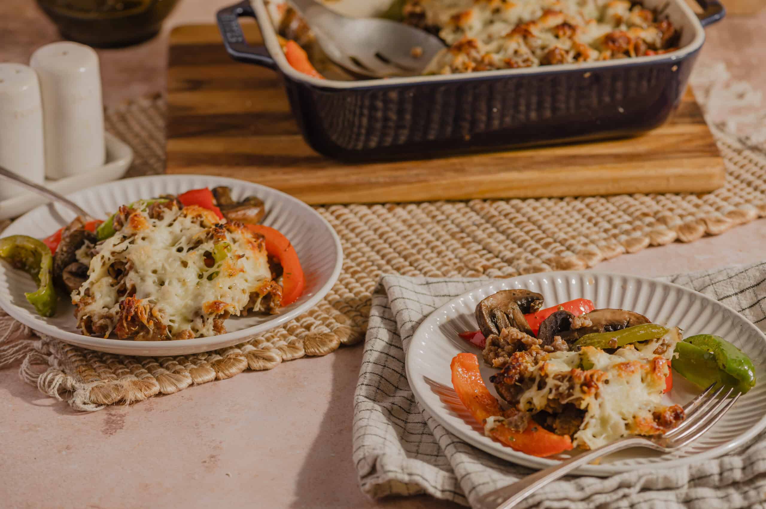 Cheesy lasagna baked dish with layers of melted cheese, seasoned ground meat, and fresh vegetables, served on white plates with colorful peppers and mushrooms, wooden cutting board and baking dish in the background.