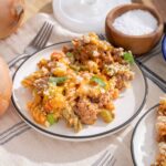 Savory ground beef and vegetable baked pasta casserole served on a white plate with shredded cheese and green onions, surrounded by onions, pasta, and rustic tabletop setting.