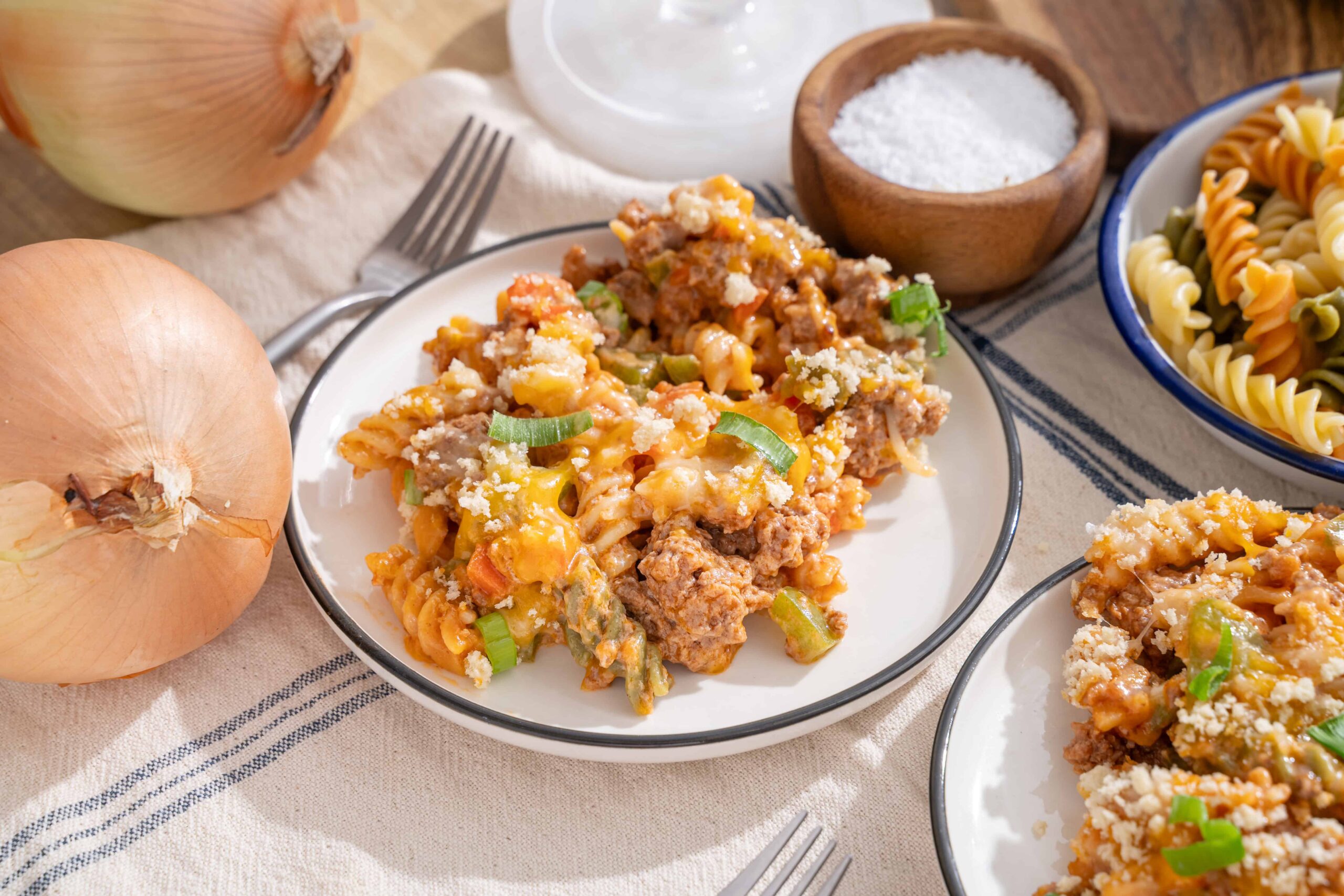 Savory ground beef and vegetable baked pasta casserole served on a white plate with shredded cheese and green onions, surrounded by onions, pasta, and rustic tabletop setting.