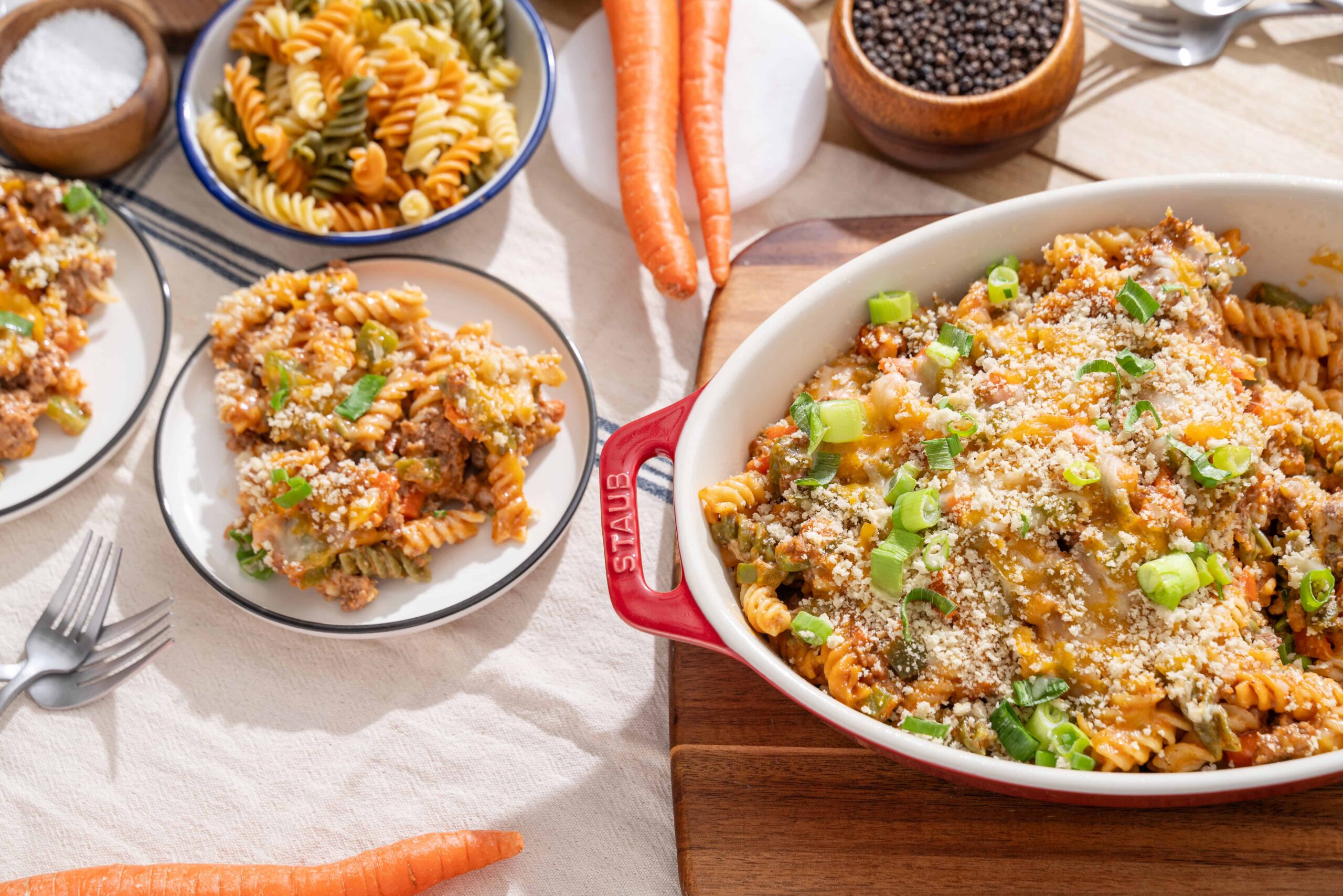 Creamy baked pasta casserole with ground beef, vegetables, and cheese garnished with green onions on a white tablecloth, surrounded by pasta bowls, carrots, and spices.
