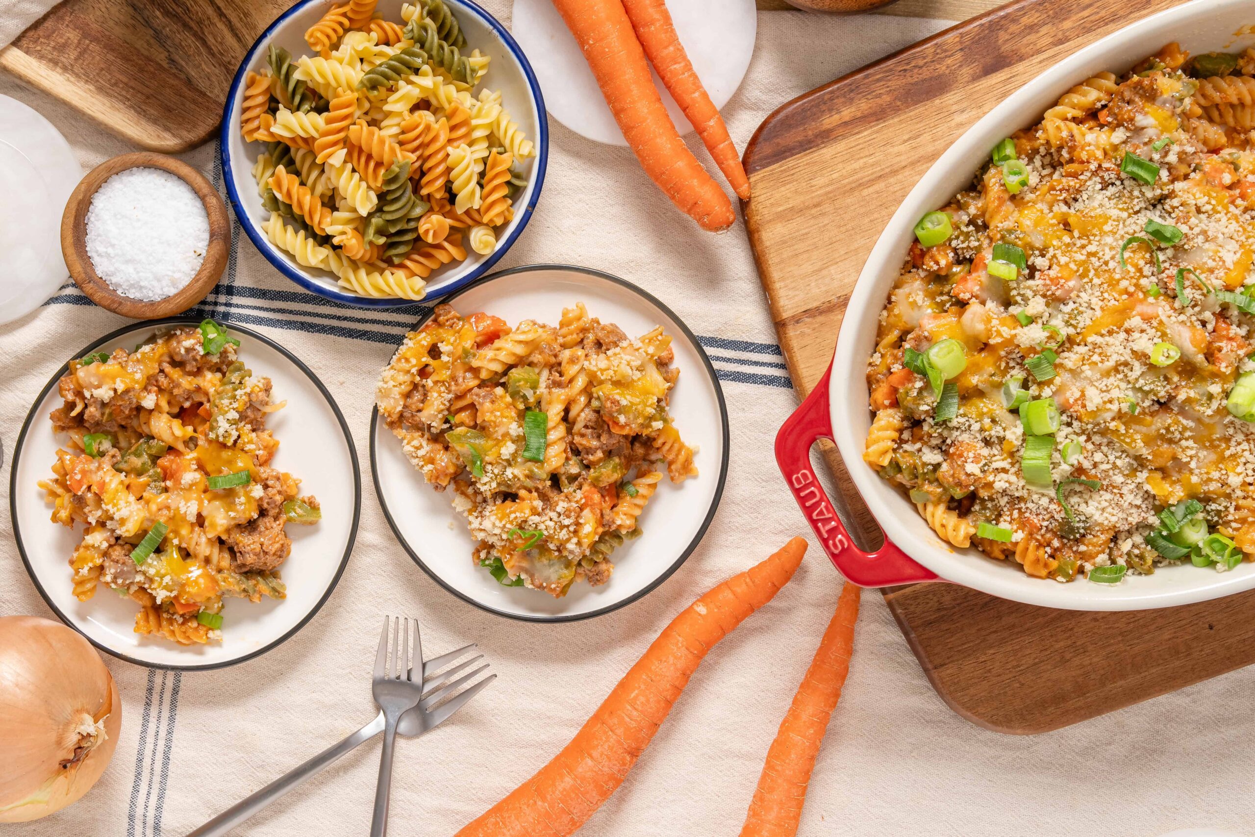 Creamy baked pasta casserole with ground beef, shredded cheese, and green onions, served in a white baking dish with colorful tri-color rotini pasta, fresh carrots, and onions on a rustic kitchen table.