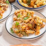 Baked rotini pasta with ground beef, cheese, and vegetables served on a white plate, surrounded by additional pasta dishes and fresh carrots.