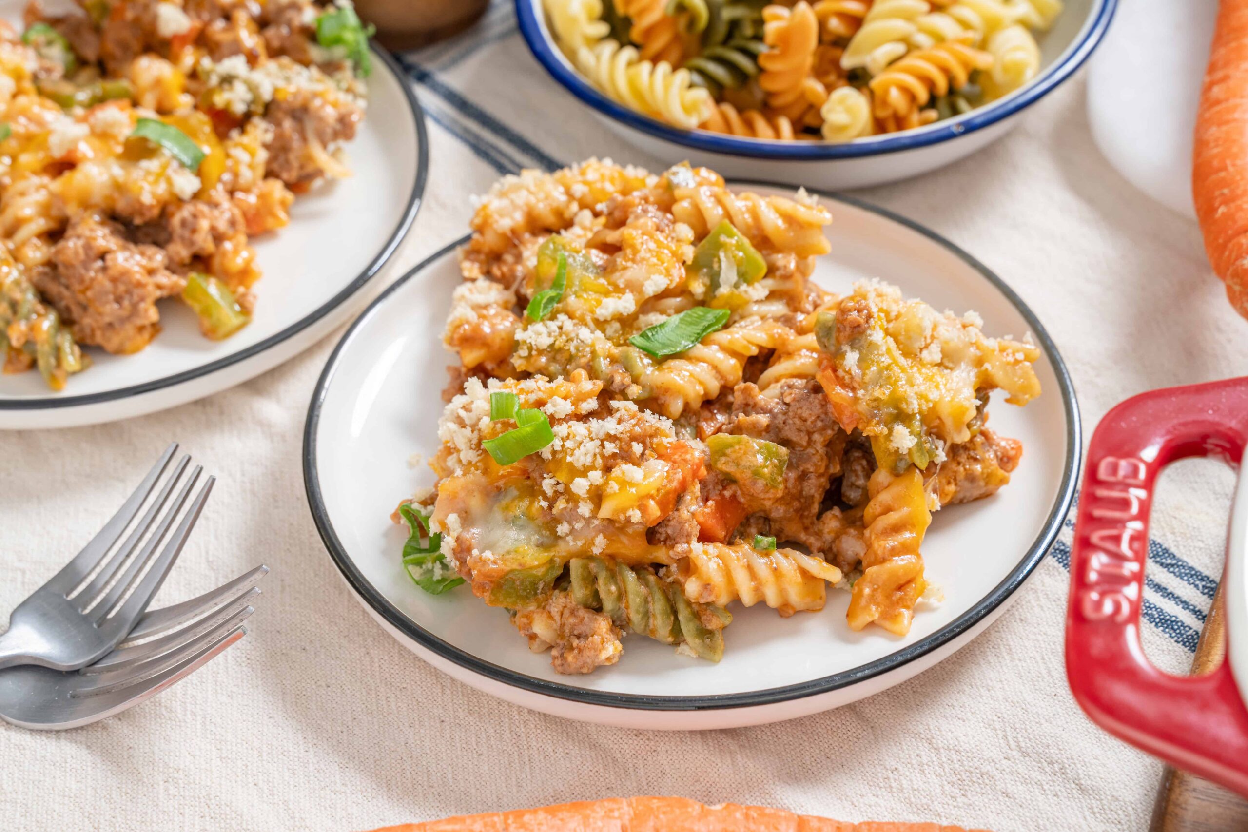 Baked rotini pasta with ground beef, cheese, and vegetables served on a white plate, surrounded by additional pasta dishes and fresh carrots.