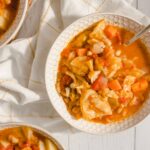 Savory vegetable soup with cabbage, carrots, and onions in a white patterned bowl, served hot with a silver spoon, on a white wooden surface with a white and gold-striped cloth, featuring fresh homemade comfort food for winter or healthy eating options.