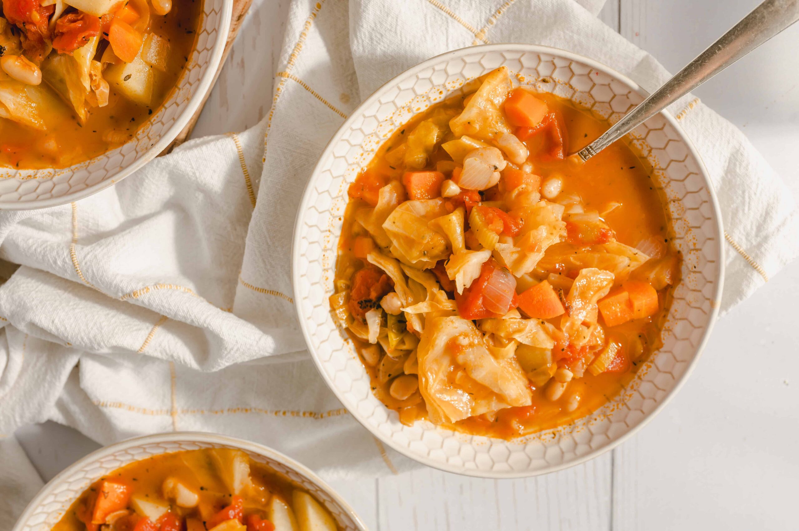 Savory vegetable soup with cabbage, carrots, and onions in a white patterned bowl, served hot with a silver spoon, on a white wooden surface with a white and gold-striped cloth, featuring fresh homemade comfort food for winter or healthy eating options.