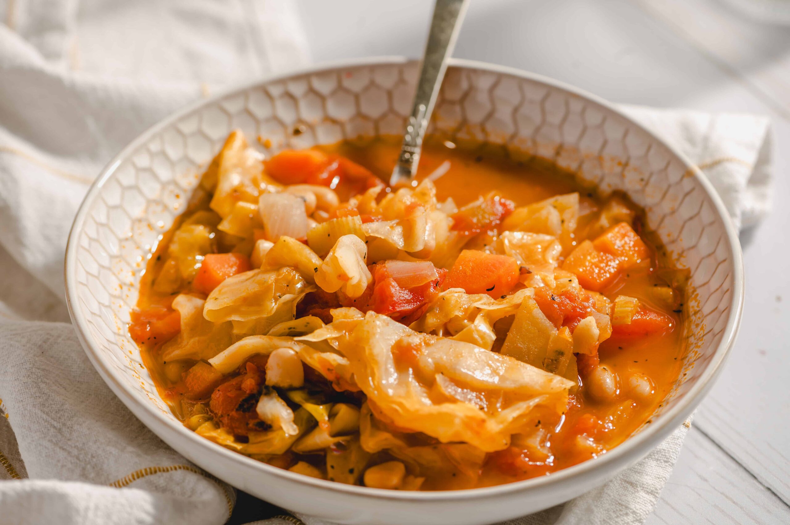 Stew with vegetables and chicken in a white bowl, garnished with herbs, on a white wooden surface with a cloth napkin in the background.