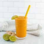 Fresh mango and lime smoothie garnished with lime wedge and served with ginger on a white surface. Perfect for a healthy, refreshing drink recipe.