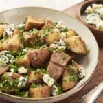 Golden roasted potatoes topped with crumbled blue cheese and chopped fresh chives, served in a rustic bowl for a hearty and flavorful side dish or appetizer.