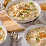 Creamy chicken vegetable soup with shredded chicken, carrots, green beans, and peas in a white bowl on a wooden cutting board and white table.