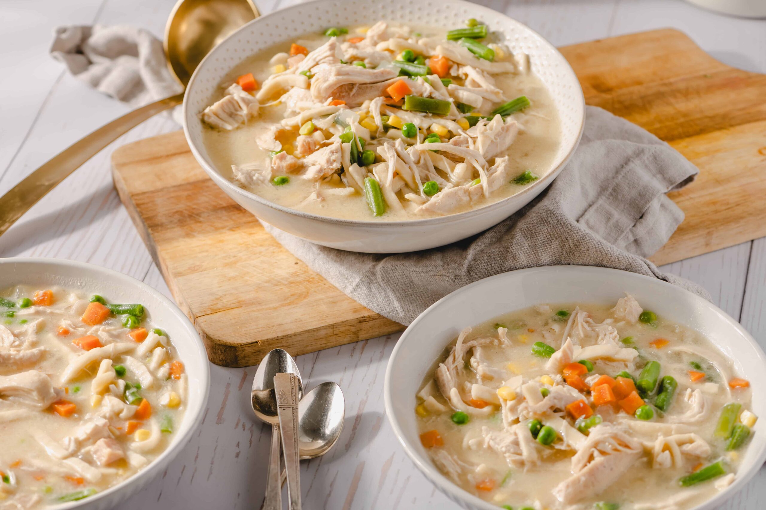 Creamy chicken vegetable soup with shredded chicken, carrots, green beans, and peas in a white bowl on a wooden cutting board and white table.