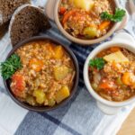 Hearty vegetable and ground meat soup served in small bowls, topped with parsley, accompanied by sliced dark bread on a checkered napkin. Ideal for comforting meals and homemade recipes.