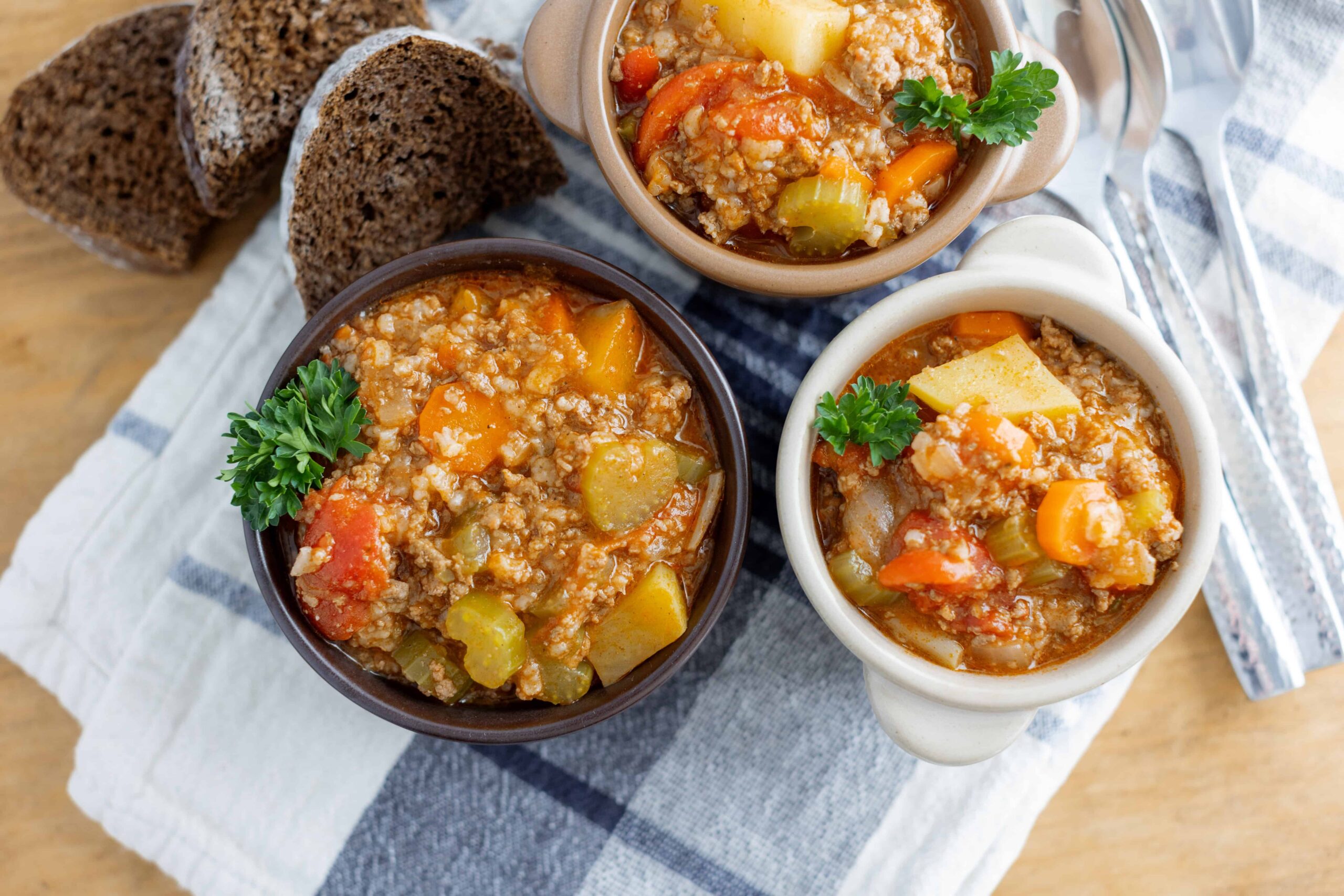 Hearty vegetable and ground meat soup served in small bowls, topped with parsley, accompanied by sliced dark bread on a checkered napkin. Ideal for comforting meals and homemade recipes.