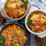 Hearty vegetable and ground meat soup served in small bowls with fresh parsley, accompanied by slices of dark rye bread on a wooden surface.