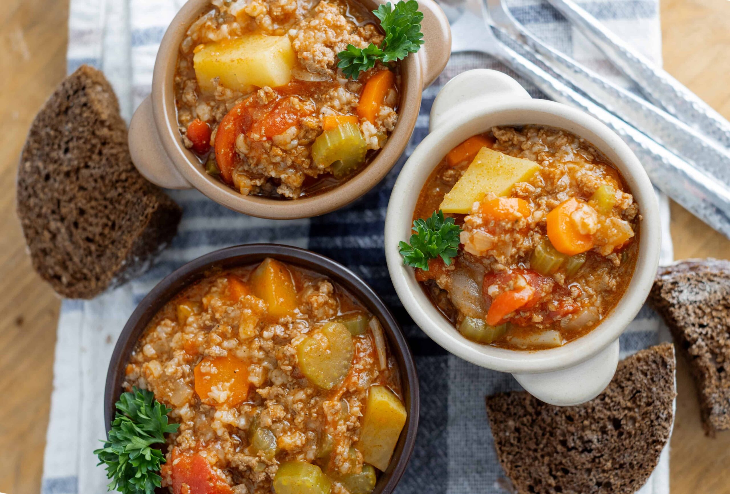 Hearty vegetable and ground meat soup served in small bowls with fresh parsley, accompanied by slices of dark rye bread on a wooden surface.