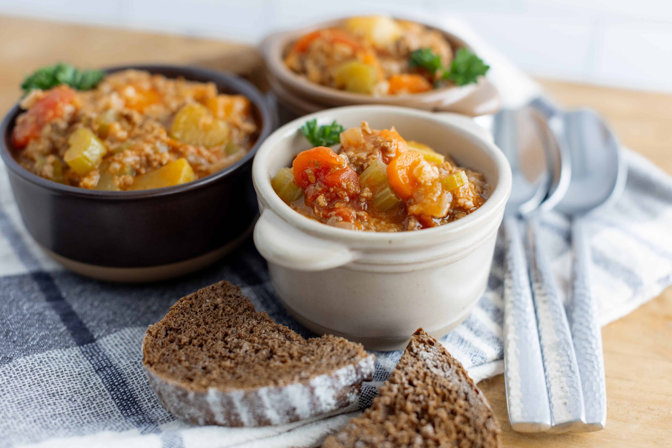 Creamy vegetable and ground meat stew served in small ceramic bowls with slices of rye bread, accompanied by silver spoons on a striped cloth.