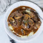 Savory mushroom and beef stew served over creamy mashed potatoes, topped with fresh herbs, in a white bowl, perfect for hearty comfort food meals.