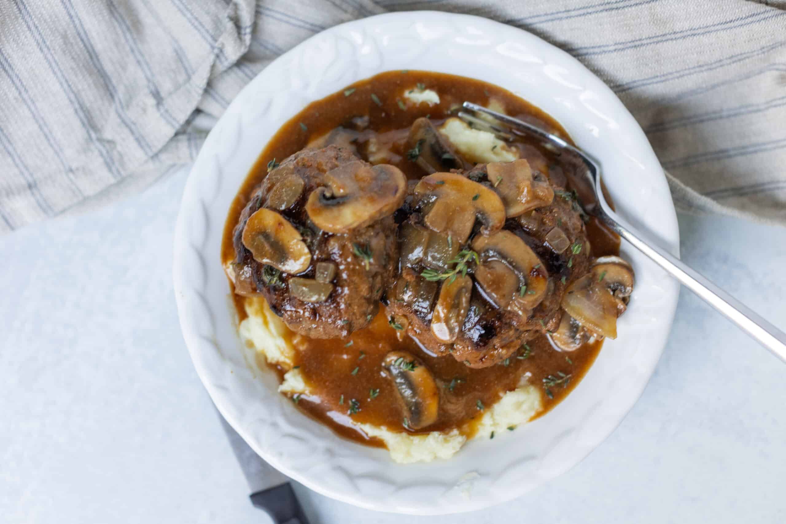 Savory mushroom and beef stew served over creamy mashed potatoes, topped with fresh herbs, in a white bowl, perfect for hearty comfort food meals.