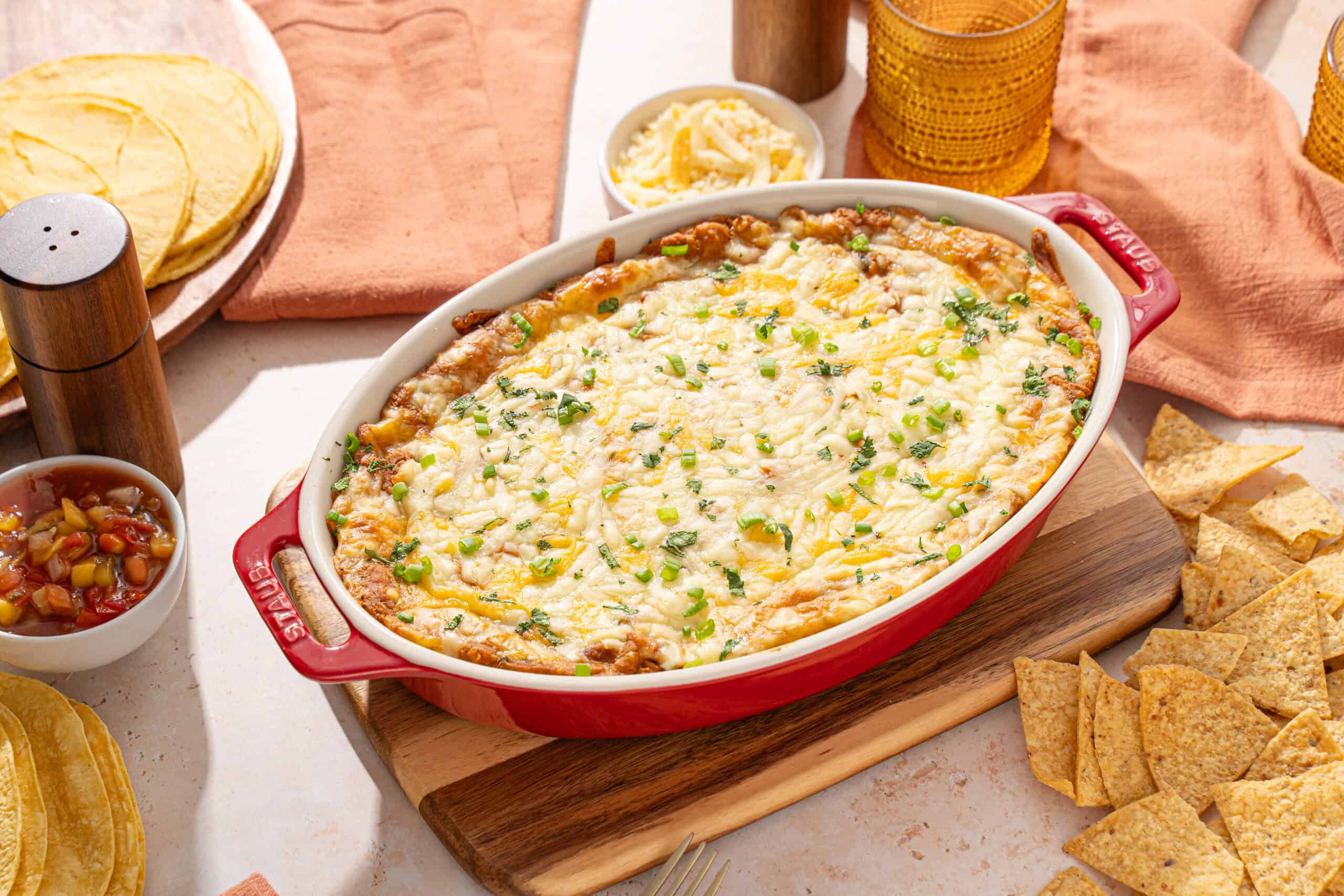 Creamy baked cheesy Mexican casserole topped with melted cheese and chopped green onions, served with tortilla chips, salsa, and fresh shredded cheese for a delicious comfort food meal.
