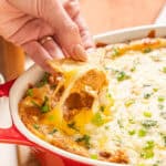Cheesy baked nachos in a red cast iron skillet with melted cheese, green onions, and seasoned ground meat, served alongside salsa and garnished with fresh herbs.