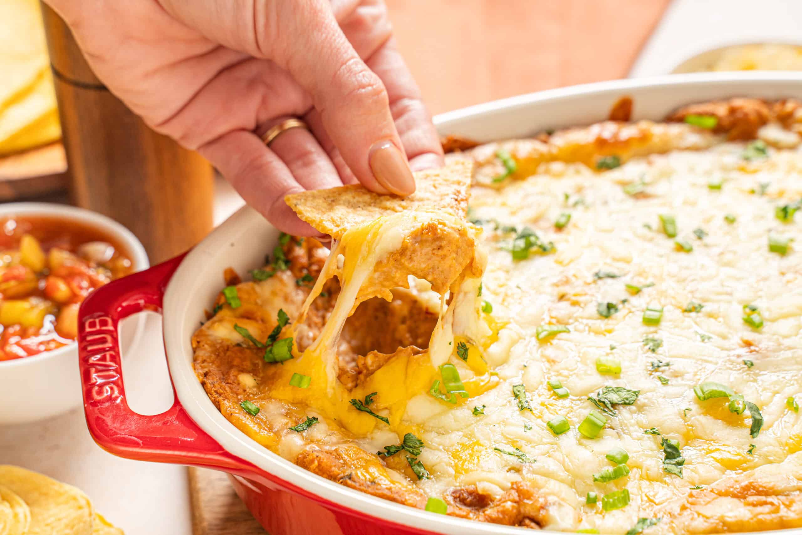 Cheesy baked nachos in a red cast iron skillet with melted cheese, green onions, and seasoned ground meat, served alongside salsa and garnished with fresh herbs.