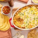 Creamy baked Mexican lasagna with melted cheese, seasoned ground beef, and green onions served in a ceramic dish. Accompanied by tortilla chips and salsa, perfect for flavorful Mexican-inspired dinner.