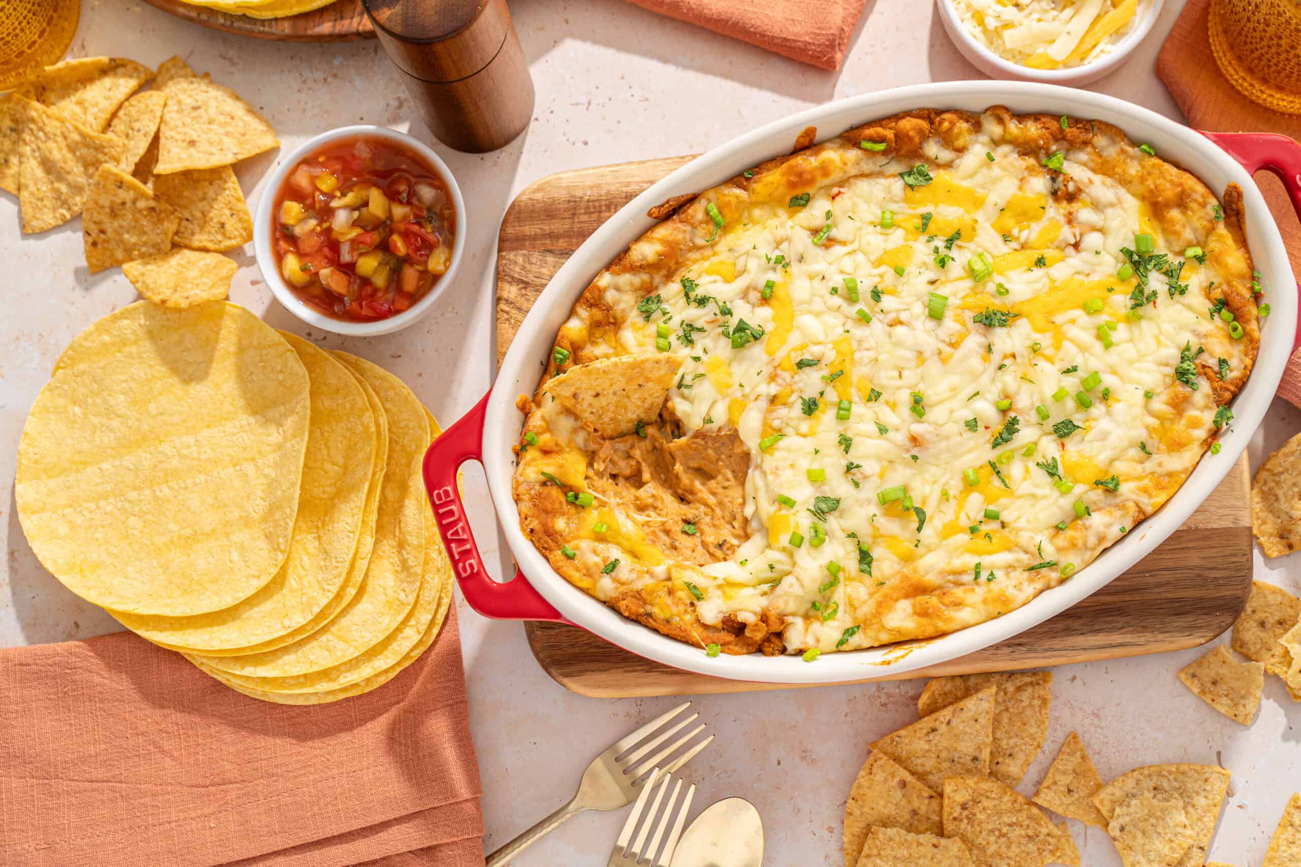 Creamy baked Mexican lasagna with melted cheese, seasoned ground beef, and green onions served in a ceramic dish. Accompanied by tortilla chips and salsa, perfect for flavorful Mexican-inspired dinner.