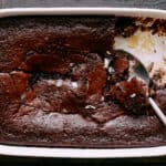 Rich, fudgy chocolate brownie with a cracked top in a white baking dish on a dark wooden surface. A spoon scoops out a piece, revealing a moist, dense interior. Perfect for dessert or chocolate cravings.