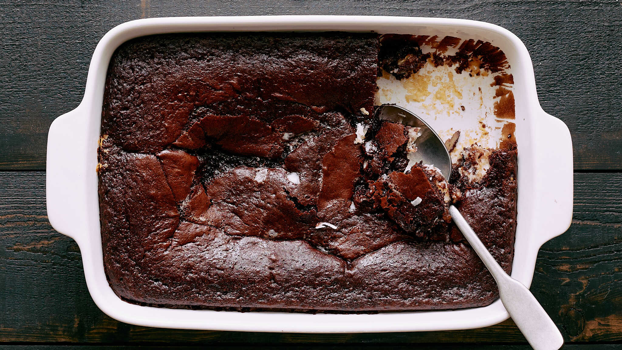 Rich, fudgy chocolate brownie with a cracked top in a white baking dish on a dark wooden surface. A spoon scoops out a piece, revealing a moist, dense interior. Perfect for dessert or chocolate cravings.